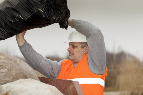 A male waste management worker wearing a white safety helmet and an orange high-visibility vest with reflective stripes is seen lifting or loading a large black garbage bag during outdoor rubbish removal. The worker's expression is focused as he handles the bag, which appears to be made of thick, durable plastic. The background shows an overcast sky with a blurred landscape of grass and possibly a construction or waste disposal site, indicating an environment related to commercial or industrial rubbish clearance. The worker is positioned on a paved or dirt surface, and his actions suggest he is engaged in a rubbish collection or clearance activity. The equipment and clothing, along with the environment, align with professional waste collection services, such as those offered by Office Clearance Forest Gate, in the London E7 postcode area.
