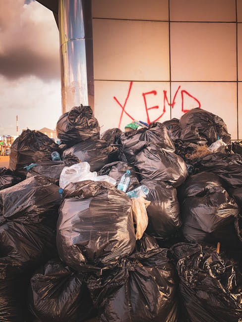 A large pile of black plastic rubbish bags, some tied and others loosely closed, stacked against a modern building with a tiled exterior wall. The bags appear full and are placed on a paved surface, w
