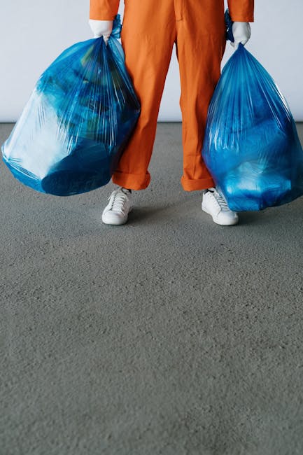 A large pile of black plastic rubbish bags, some tied and others loosely closed, stacked against a modern building with a tiled exterior wall. The bags appear full and are placed on a paved surface, w