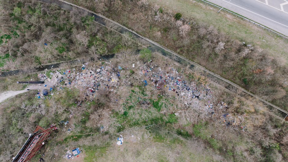 An aerial view of a large, cluttered, and unmanaged refuse pile situated on a patch of open land adjacent to a paved road and a railway line. The rubbish consists of various materials including plasti
