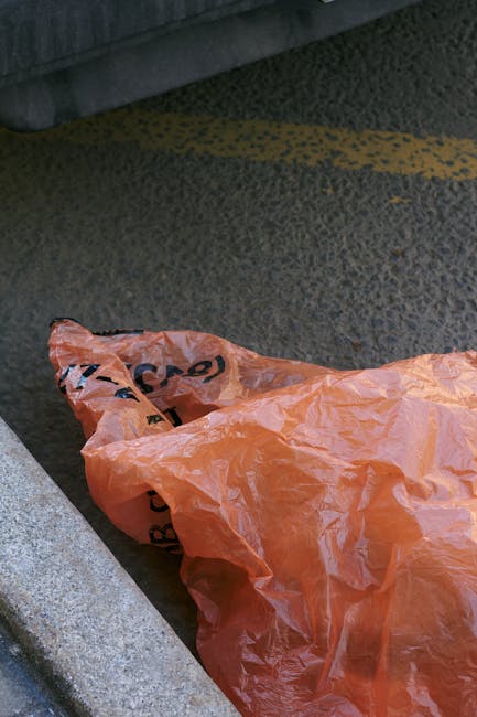 An aerial view of a large, cluttered, and unmanaged refuse pile situated on a patch of open land adjacent to a paved road and a railway line. The rubbish consists of various materials including plasti