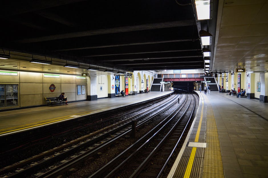 A view of an underground train station platform illuminated by overhead lighting, with multiple train tracks running parallel along the left side. The platform surface is made of concrete with tactile