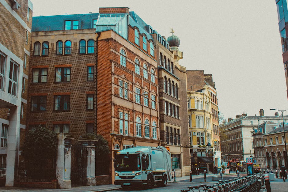 The image shows a large collection of mixed waste and rubbish overflowing from multiple public trash bins and containers situated on a paved urban street outside a row of small retail shops. The debri