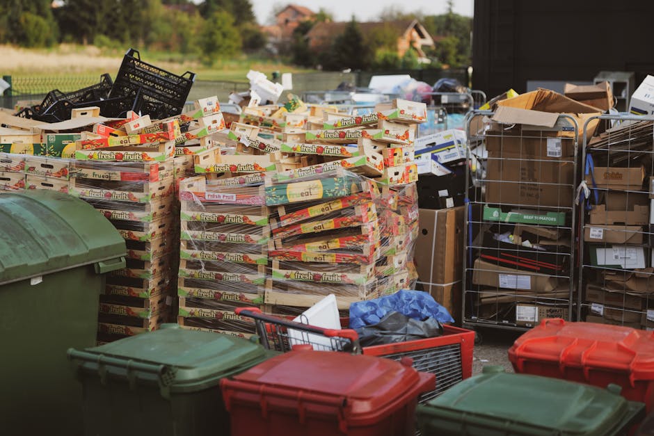 An outdoor scene showing a large quantity of discarded cardboard boxes, primarily from fresh fruit packaging, stacked in front of a fence. The boxes are colorful with red, green, and yellow branding,